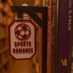 Op de afbeelding: Een zwart en rood 3D-geprint boekensteun met een voetbalontwerp en de tekst "SPORTS ROMANCE". De boekensteun staat tussen boeken op een plank, wat wijst op een thema van sportromans. Het ontwerp is rood en wit.