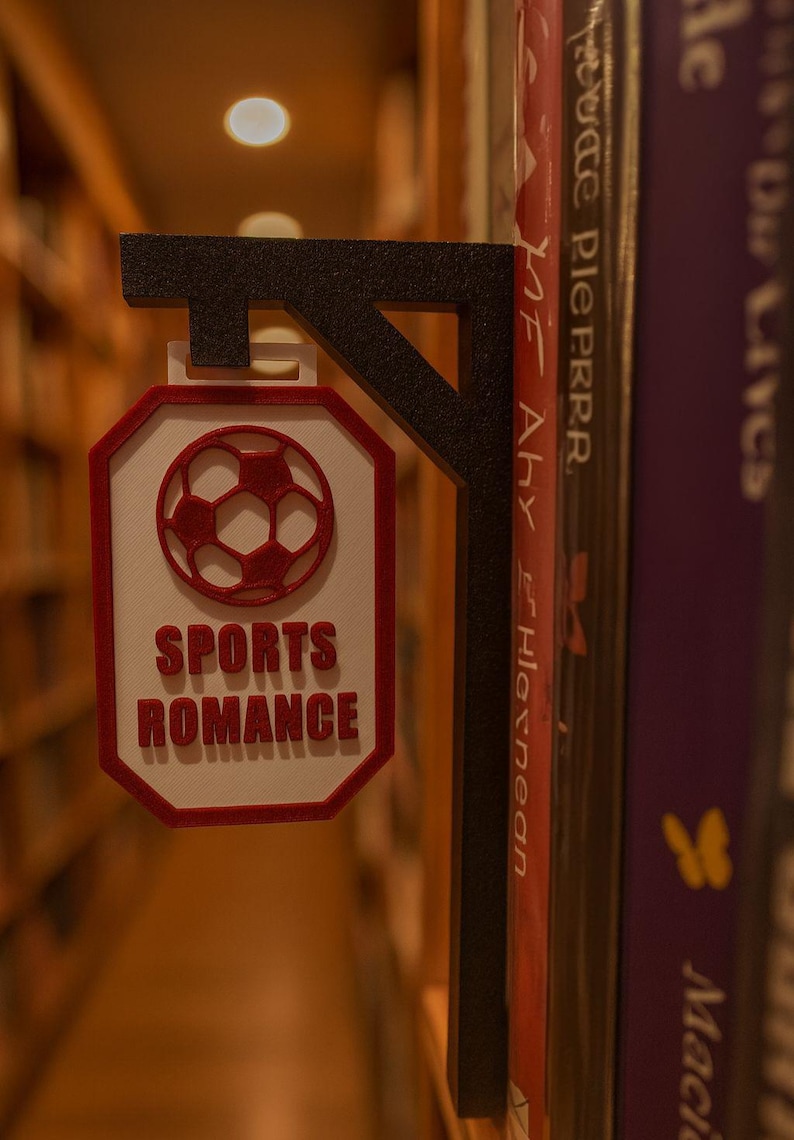 Op de afbeelding: Een zwart en rood 3D-geprint boekensteun met een voetbalontwerp en de tekst "SPORTS ROMANCE". De boekensteun staat tussen boeken op een plank, wat wijst op een thema van sportromans. Het ontwerp is rood en wit.