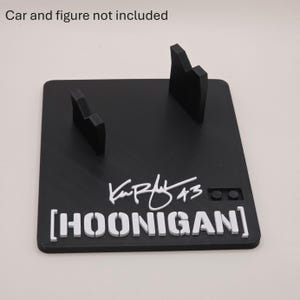 May include: A black square display stand featuring two upright supports, a white signature with the number 43, and the white text HOONIGAN in brackets. The surface includes a small two-stud connector block. The text Car and figure not included is visible.