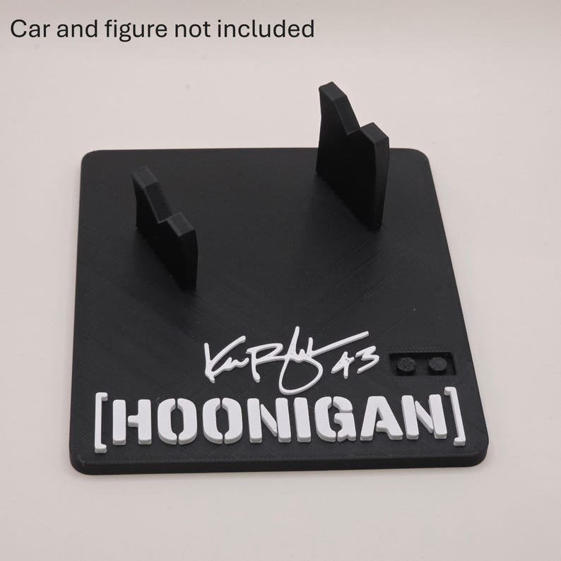 May include: A black square display stand featuring two upright supports, a white signature with the number 43, and the white text HOONIGAN in brackets. The surface includes a small two-stud connector block. The text Car and figure not included is visible.