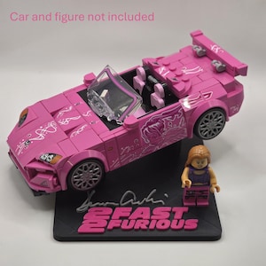 May include: A pink brick-built convertible sports car with white graphic decals, a clear windshield, and grey wheels. A small plastic figure with long blonde hair and a purple outfit stands on a black display base labeled 2 FAST 2 FURIOUS.