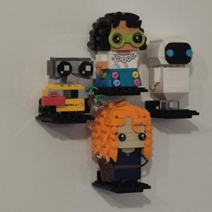 Op de afbeelding: Een verzameling van vijf LEGO minifiguren die op planken worden tentoongesteld. De minifiguren omvatten WALL-E, een vrouwelijke minifiguur met bruin haar en groene brillen, een witte robot met blauwe ogen en een vrouwelijke minifiguur met oranje haar.