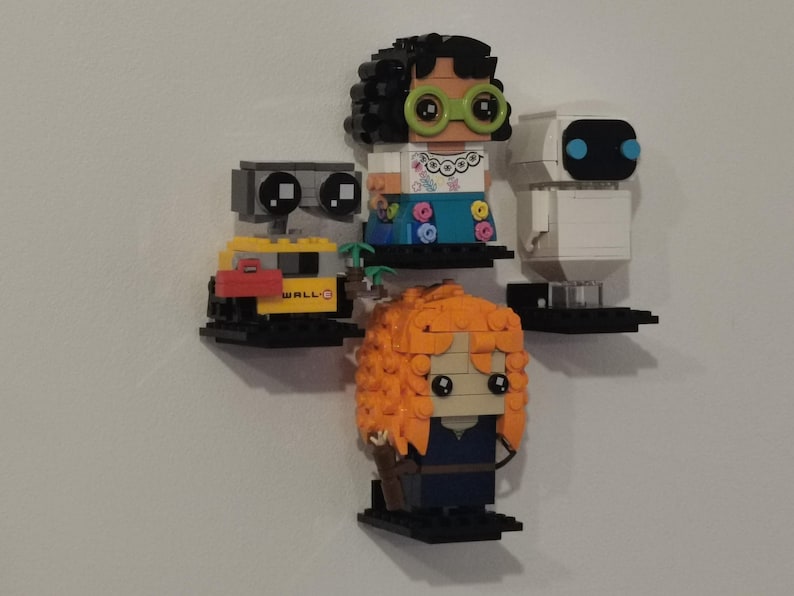 Op de afbeelding: Een verzameling van vijf LEGO minifiguren die op planken worden tentoongesteld. De minifiguren omvatten WALL-E, een vrouwelijke minifiguur met bruin haar en groene brillen, een witte robot met blauwe ogen en een vrouwelijke minifiguur met oranje haar.