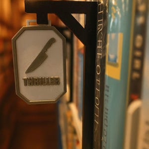 Op de afbeelding: Een zwart metalen boekenplankbord met een wit en goudkleurig achthoekig bord met de tekst "THRILLER" en een mesafbeelding. Het bord is bevestigd aan een boekenplank met verschillende boeken.