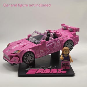May include: A black display stand featuring the text 2 FAST 2 FURIOUS in pink lettering. The stand includes a mounting bracket designed to hold a pink toy car and a small plastic figure, with the text Car and figure not included visible above.