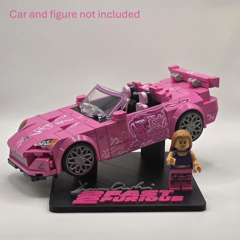 May include: A black display stand featuring the text 2 FAST 2 FURIOUS in pink lettering. The stand includes a mounting bracket designed to hold a pink toy car and a small plastic figure, with the text Car and figure not included visible above.