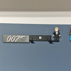 Puede incluir: Un soporte de pared negro con el n&uacute;mero "007" y un s&iacute;mbolo de pistola impreso en &eacute;l. Una minifigura de Lego de un hombre con esmoquin est&aacute; de pie en el soporte.