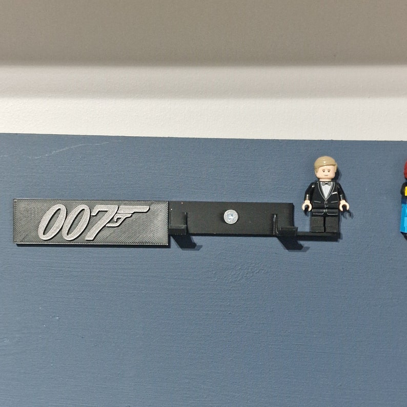 Puede incluir: Un soporte de pared negro con el n&uacute;mero "007" y un s&iacute;mbolo de pistola impreso en &eacute;l. Una minifigura de Lego de un hombre con esmoquin est&aacute; de pie en el soporte.