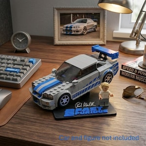 May include: A silver and blue brick-built sports car model with a rear spoiler, displayed on a black stand featuring the text 2 Fast 2 Furious and a signature. A small plastic figure stands next to the car on the base. The text Car and figure not included is visible at the bottom.