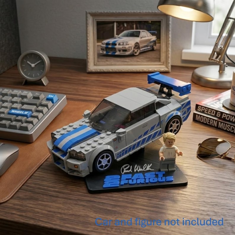 May include: A silver and blue brick-built sports car model with a rear spoiler, displayed on a black stand featuring the text 2 Fast 2 Furious and a signature. A small plastic figure stands next to the car on the base. The text Car and figure not included is visible at the bottom.