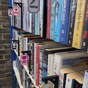 Op de afbeelding: Een boekenplank met diverse boeken en genresignalen. De borden zijn zwart met witte en gekleurde accenten, die genres aangeven zoals "Thriller" met een mes-icoon, "Romance" met een hart en "Fantasy" met een vleermuis.