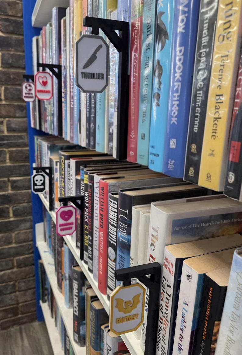 Op de afbeelding: Een boekenplank met diverse boeken en genresignalen. De borden zijn zwart met witte en gekleurde accenten, die genres aangeven zoals "Thriller" met een mes-icoon, "Romance" met een hart en "Fantasy" met een vleermuis.
