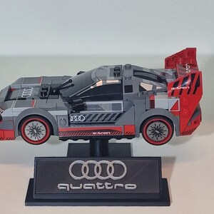May include: A gray and red Lego Audi e-tron quattro race car model with a black display stand featuring the Audi logo and the word "quattro".