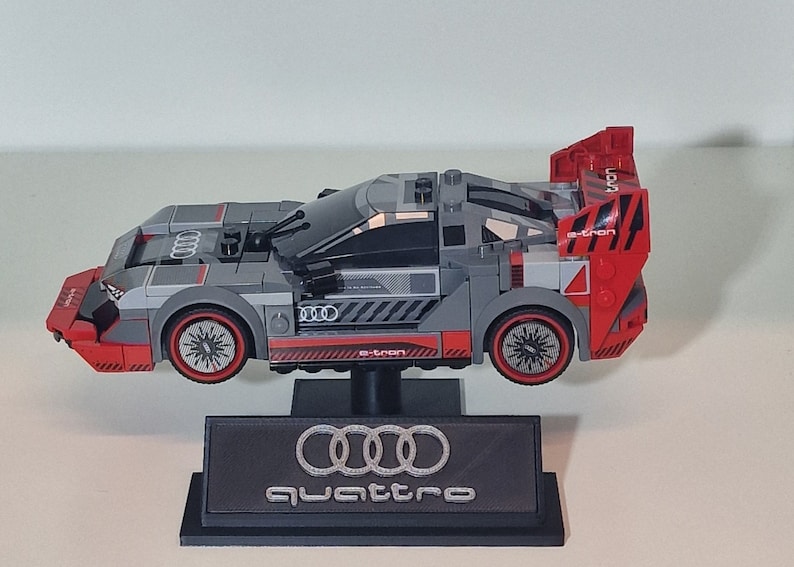 May include: A gray and red Lego Audi e-tron quattro race car model with a black display stand featuring the Audi logo and the word "quattro".