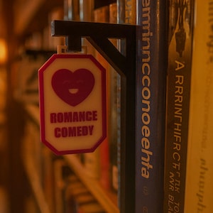 Op de afbeelding: Een bord voor het genre Romantische Komedie, met een hartvormige afbeelding en de woorden "ROMANCE COMEDY" in het rood. Het bord is bevestigd aan een zwarte beugel, tussen een boekenplank.