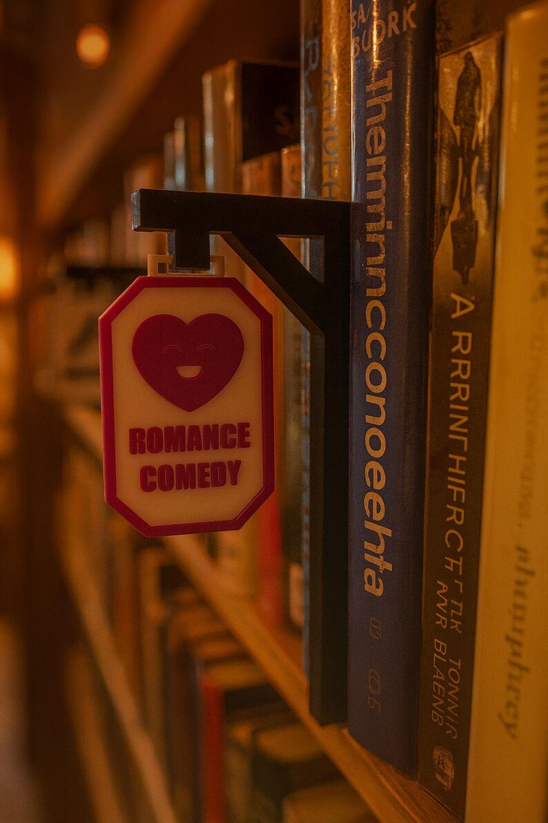 Op de afbeelding: Een bord voor het genre Romantische Komedie, met een hartvormige afbeelding en de woorden "ROMANCE COMEDY" in het rood. Het bord is bevestigd aan een zwarte beugel, tussen een boekenplank.