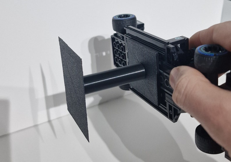 May include: A black Lego car model with blue wheels is mounted on a black stand with a square base. The stand is made of a 3D printed material.