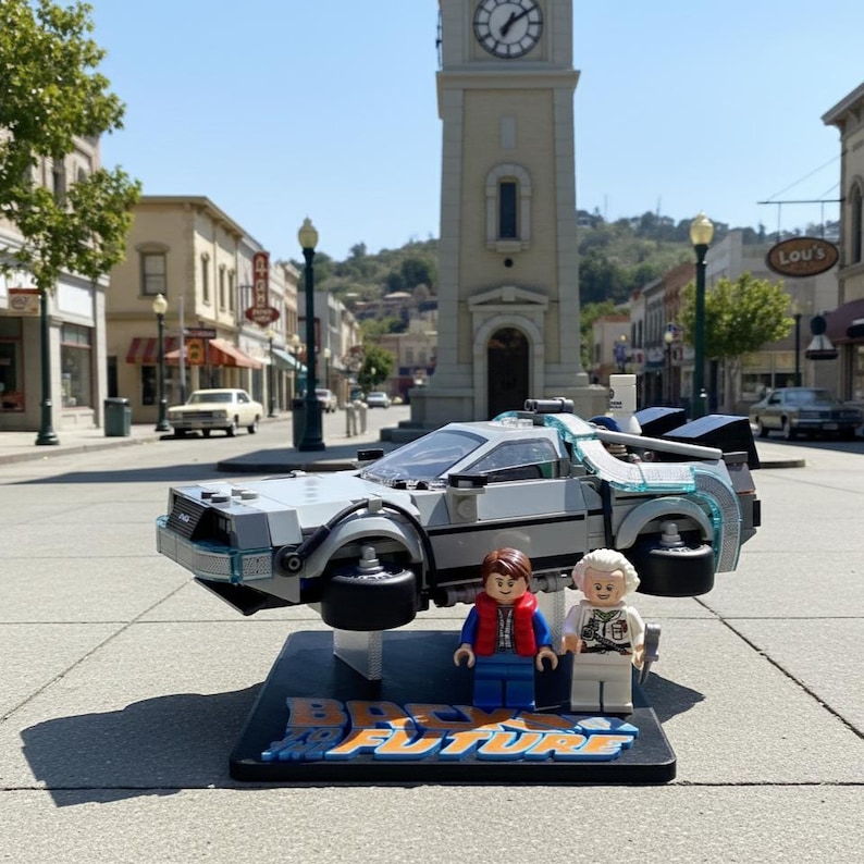 May include: A Lego DeLorean time machine model from the movie Back to the Future, with two Lego figures. The car is gray with blue accents and hovers above a black base with the movie title. The background shows a town square.