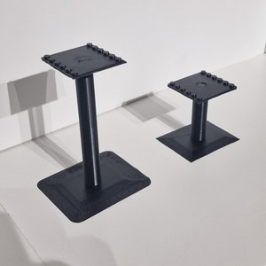 May include: Two black 3D printed stands with square bases and a cylindrical post. The larger stand is taller than the smaller stand.