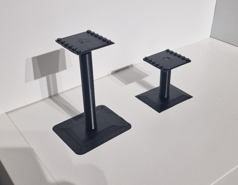 May include: Two black 3D printed stands with square bases and a cylindrical post. The larger stand is taller than the smaller stand.