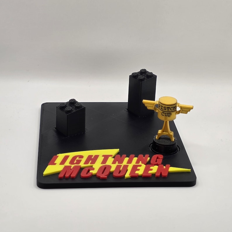 Puede incluir: Una base negra con las palabras "Lightning McQueen" en rojo y amarillo. La base tiene dos estructuras de bloques negros y un trofeo de la Copa Pist&oacute;n dorado. Un objeto de colecci&oacute;n para los fans de la pel&iacute;cula Cars.