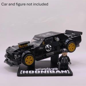 May include: A black plastic building block model of a Ford Mustang car with gold wheels, Hoonigan branding, and a matching minifigure standing on a black display base with the text HOONIGAN and a signature. Car and figure not included.