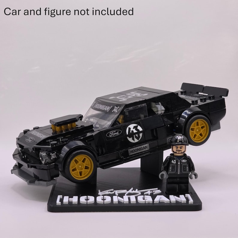 May include: A black plastic building block model of a Ford Mustang car with gold wheels, Hoonigan branding, and a matching minifigure standing on a black display base with the text HOONIGAN and a signature. Car and figure not included.