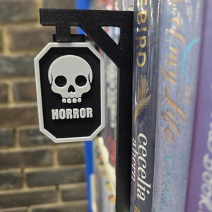 Op de afbeelding: Zwart metalen boekenplankbord met een witte schedelafbeelding en het woord "HORROR" in witte blokletters. Het bord is bevestigd aan een boekenplank met boeken.