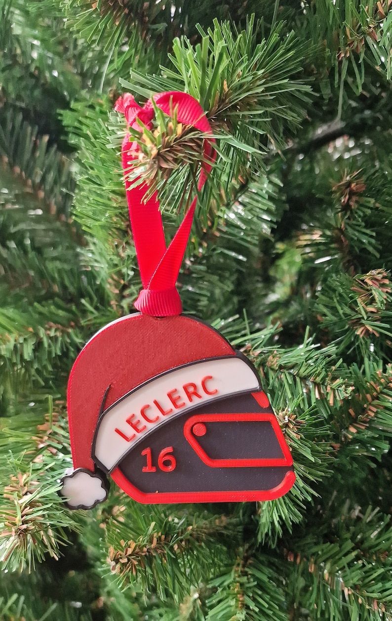 Op de afbeelding: Een kerstboomversiering in de vorm van een racehelm en een kerstmuts. De rood-zwarte helm heeft de naam "LECLERC" en het nummer "16". Een rood lint is bevestigd om op te hangen.