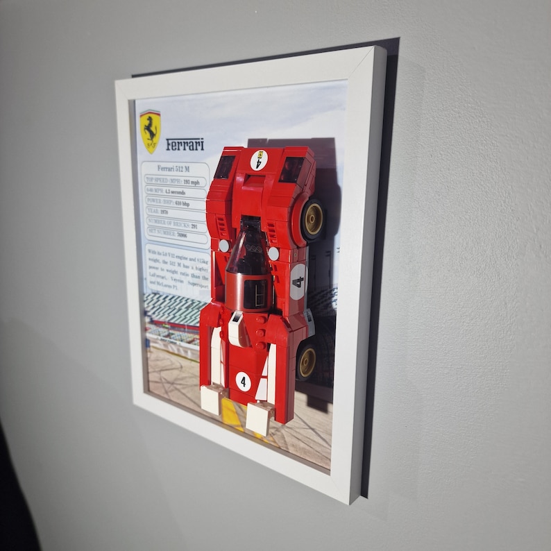 以下が含まれることがあります： おもちゃのブロックで作られた赤いフェラーリ512Mレーシングカーモデルを特徴とする額装されたアートワーク。モデルは技術仕様とフェラーリのロゴを背景に配置されています。フレームは白です。