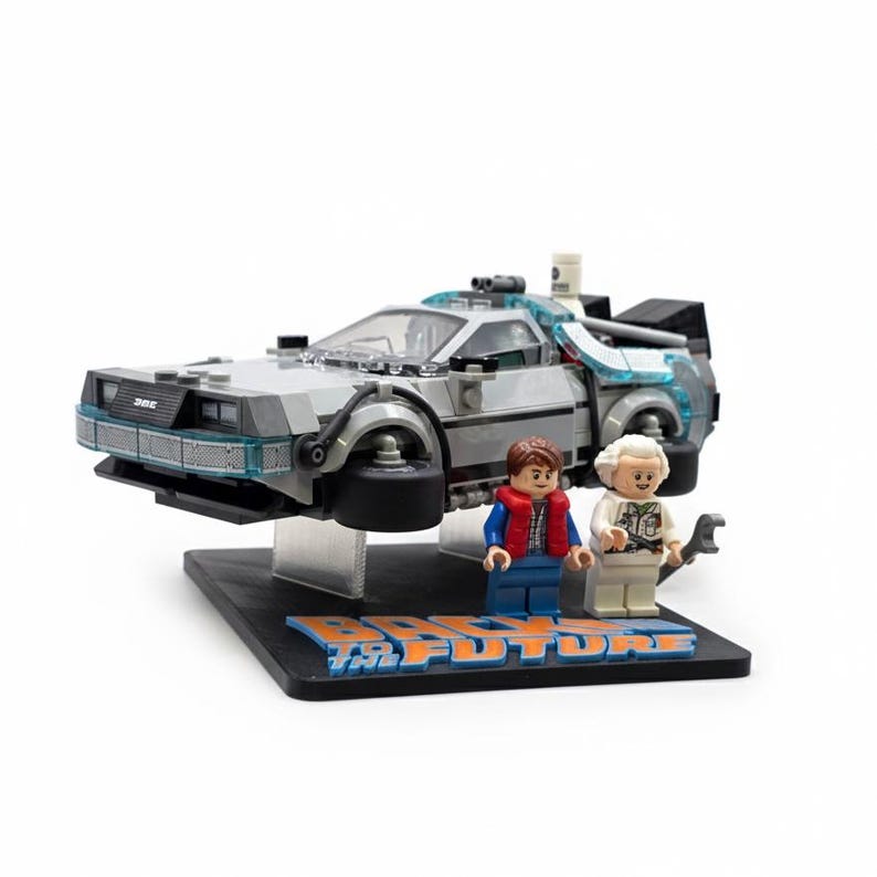 May include: A Lego model of the DeLorean time machine from Back to the Future, with two minifigures. The car is silver and gray, with blue accents. The base has the words "Back to the Future" in blue and orange.