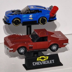 May include: Two toy Chevrolet race cars. The top car is blue with black accents and the number 5. The bottom car is red. Both cars are on black stands. The Chevrolet logo is visible on the bottom stand.