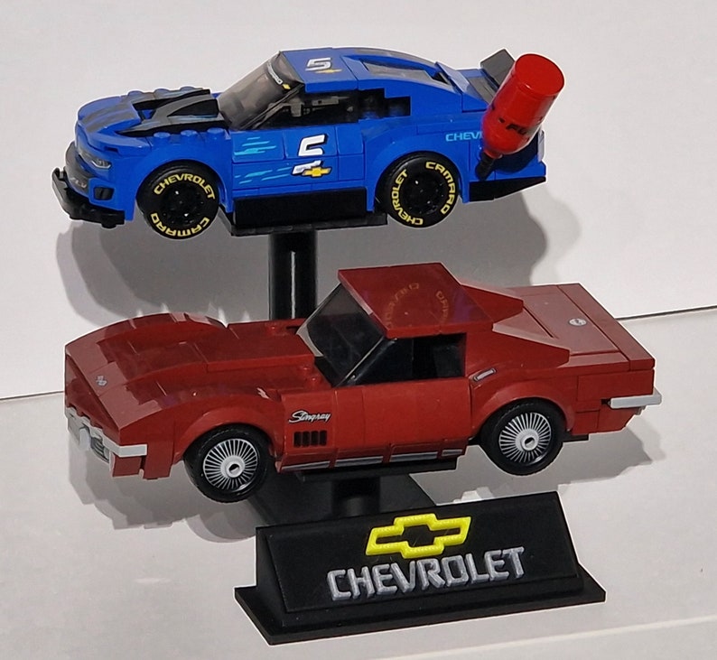 May include: Two toy Chevrolet race cars. The top car is blue with black accents and the number 5. The bottom car is red. Both cars are on black stands. The Chevrolet logo is visible on the bottom stand.