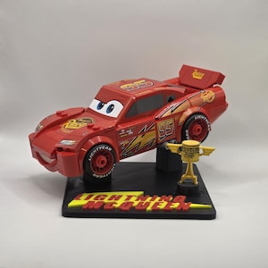 Puede incluir: Un coche de juguete rojo Lightning McQueen con el n&uacute;mero 95 en el lateral, un aler&oacute;n y ojos grandes. El coche est&aacute; sobre una base negra con las palabras "Lightning McQueen" en amarillo. Un peque&ntilde;o trofeo dorado tambi&eacute;n est&aacute; en la base.