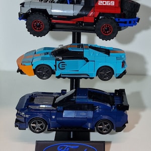 Puede incluir: Un expositor de tres niveles con un logotipo de Ford azul en la base. El expositor presenta tres veh&iacute;culos Ford diferentes: un Ford Mustang azul, un Ford GT azul claro y un Ford Raptor rojo y gris.