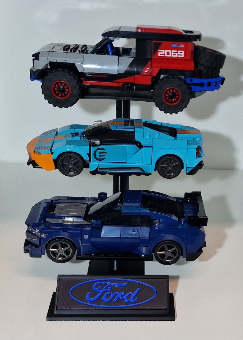 Puede incluir: Un expositor de tres niveles con un logotipo de Ford azul en la base. El expositor presenta tres veh&iacute;culos Ford diferentes: un Ford Mustang azul, un Ford GT azul claro y un Ford Raptor rojo y gris.