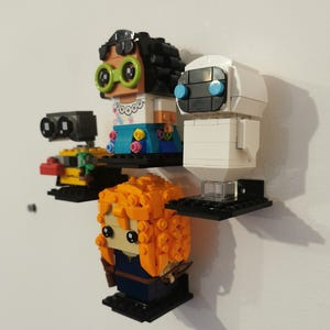 Op de afbeelding: Een set van vier Lego minifiguren die op zwarte planken worden getoond. De minifiguren omvatten een vrouwelijke figuur met bruin haar en groene brillen, een witte robot met blauwe ogen, een gele robot met zwarte ogen en een vrouwelijke figuur met oranje haar.