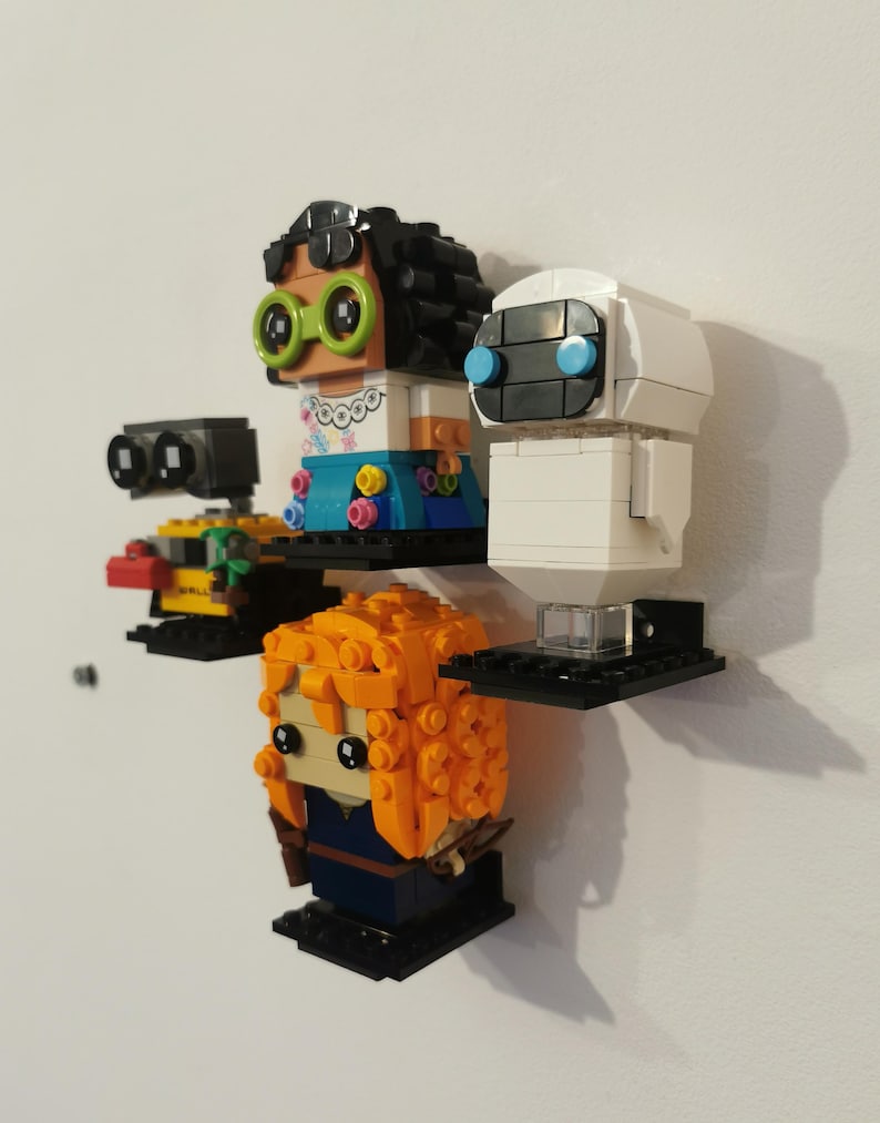Op de afbeelding: Een set van vier Lego minifiguren die op zwarte planken worden getoond. De minifiguren omvatten een vrouwelijke figuur met bruin haar en groene brillen, een witte robot met blauwe ogen, een gele robot met zwarte ogen en een vrouwelijke figuur met oranje haar.