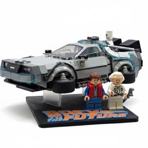 May include: A Lego DeLorean time machine model with two Lego figures. The car is gray with blue accents and black wheels. The figures are of a man in a red vest and a man in a white coat. The base reads "Back to the Future".