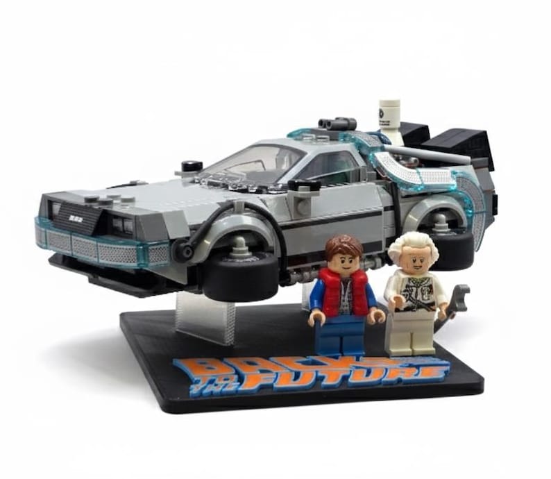 May include: A Lego DeLorean time machine model with two Lego figures. The car is gray with blue accents and black wheels. The figures are of a man in a red vest and a man in a white coat. The base reads "Back to the Future".