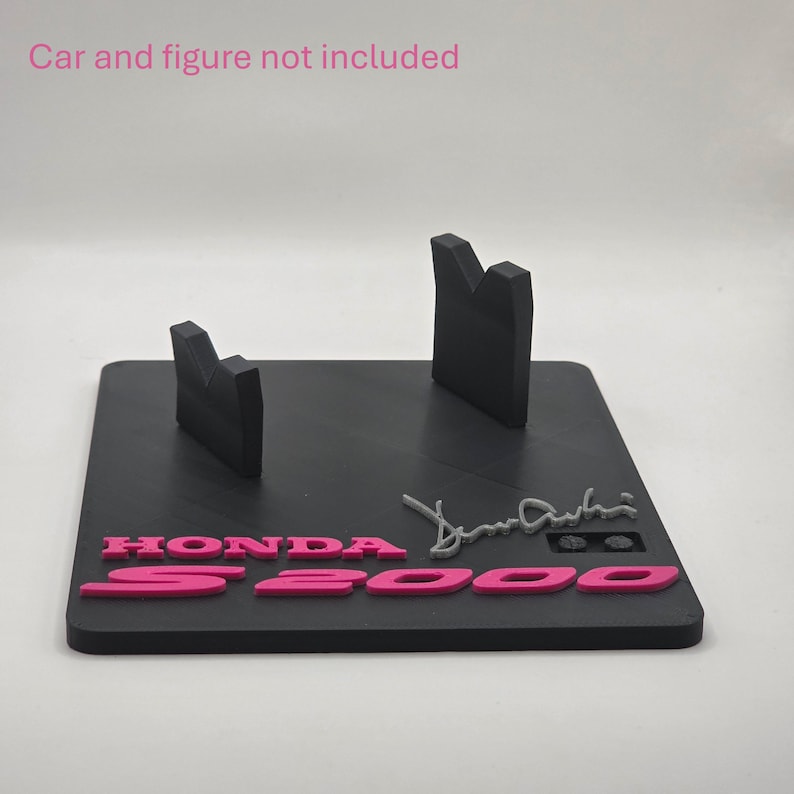 May include: A black rectangular display stand featuring two upright supports for a model vehicle. The base displays the pink text HONDA S2000 and a grey signature. The text Car and figure not included is visible at the top of the image.