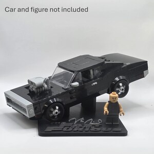 May include: A black brick-built muscle car model with a prominent silver engine block protruding from the hood, displayed on a black stand featuring the text "The Fast and the Furious". A small plastic figure stands beside the car. Car and figure not included.
