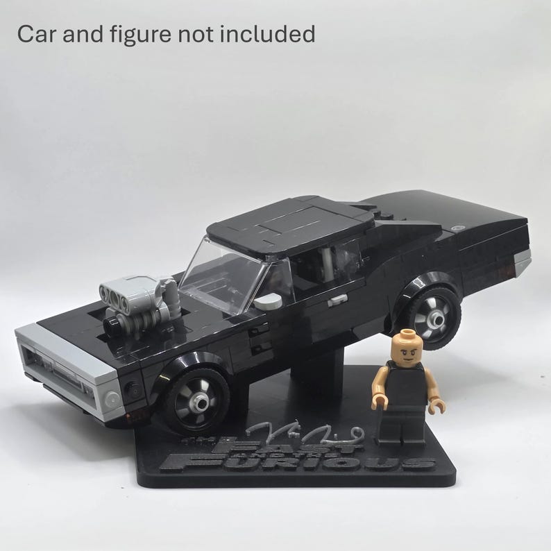 May include: A black brick-built muscle car model with a prominent silver engine block protruding from the hood, displayed on a black stand featuring the text "The Fast and the Furious". A small plastic figure stands beside the car. Car and figure not included.