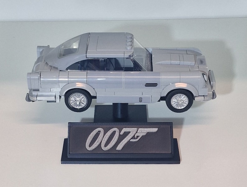 May include: A silver Lego model of the iconic Aston Martin DB5 from the James Bond films, displayed on a black stand with the 007 logo.
