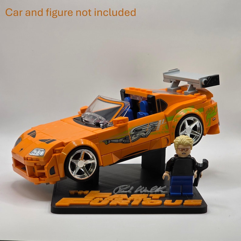 Pu&ograve; includere: Un'auto sportiva in mattoncini arancione brillante con alettone posteriore, ruote argentate e decalcomanie verdi, su una base nera con la scritta "Fast & Furious" e una statuetta bionda con una chiave inglese. Testo: Car and figure not included.