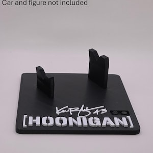 May include: A black rectangular display stand featuring two upright supports, the white text HOONIGAN, a white signature, and the number 43. The text Car and figure not included is printed above the stand. The base has a textured surface.