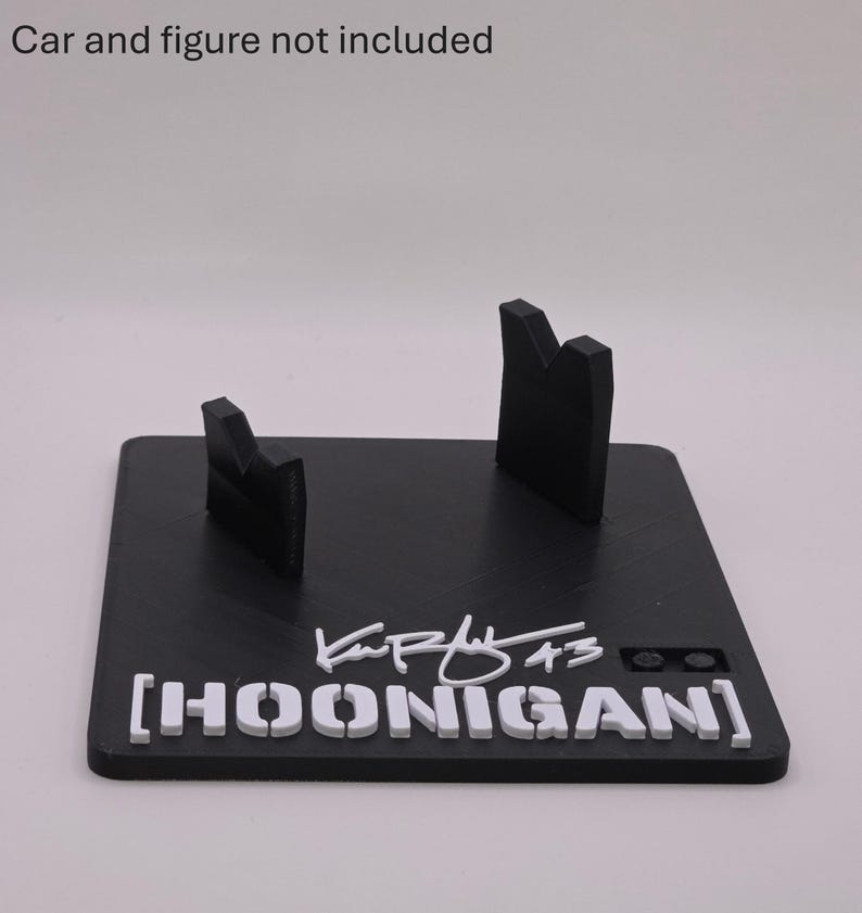 May include: A black rectangular display stand featuring two upright supports, the white text HOONIGAN, a white signature, and the number 43. The text Car and figure not included is printed above the stand. The base has a textured surface.