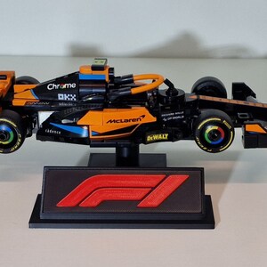 May include: A black and orange Lego McLaren Formula 1 race car with the logos "Chrome", "OKX", "McLaren", "DeWalt", and "Richard Mille" on the side. The car is on a black display stand with the Formula 1 logo.