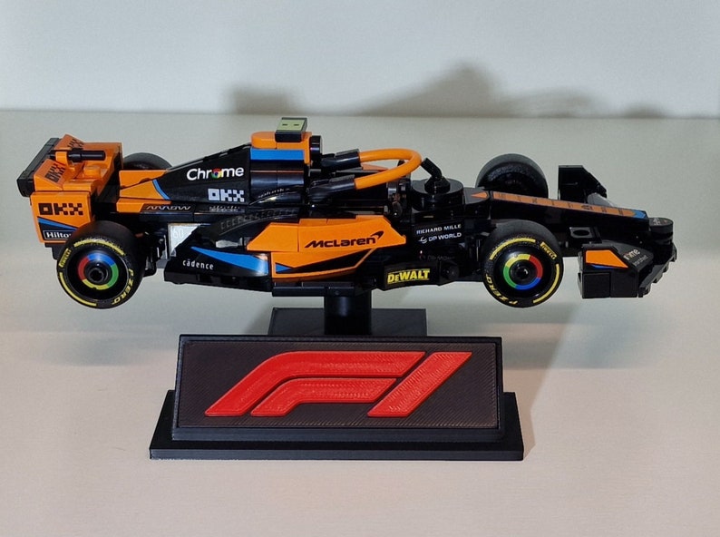 May include: A black and orange Lego McLaren Formula 1 race car with the logos "Chrome", "OKX", "McLaren", "DeWalt", and "Richard Mille" on the side. The car is on a black display stand with the Formula 1 logo.