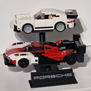 May include: Two toy Porsche race cars, one white and one red and black, are displayed on a black stand. The white car is on top, and the red and black car is below. The stand has the Porsche logo.